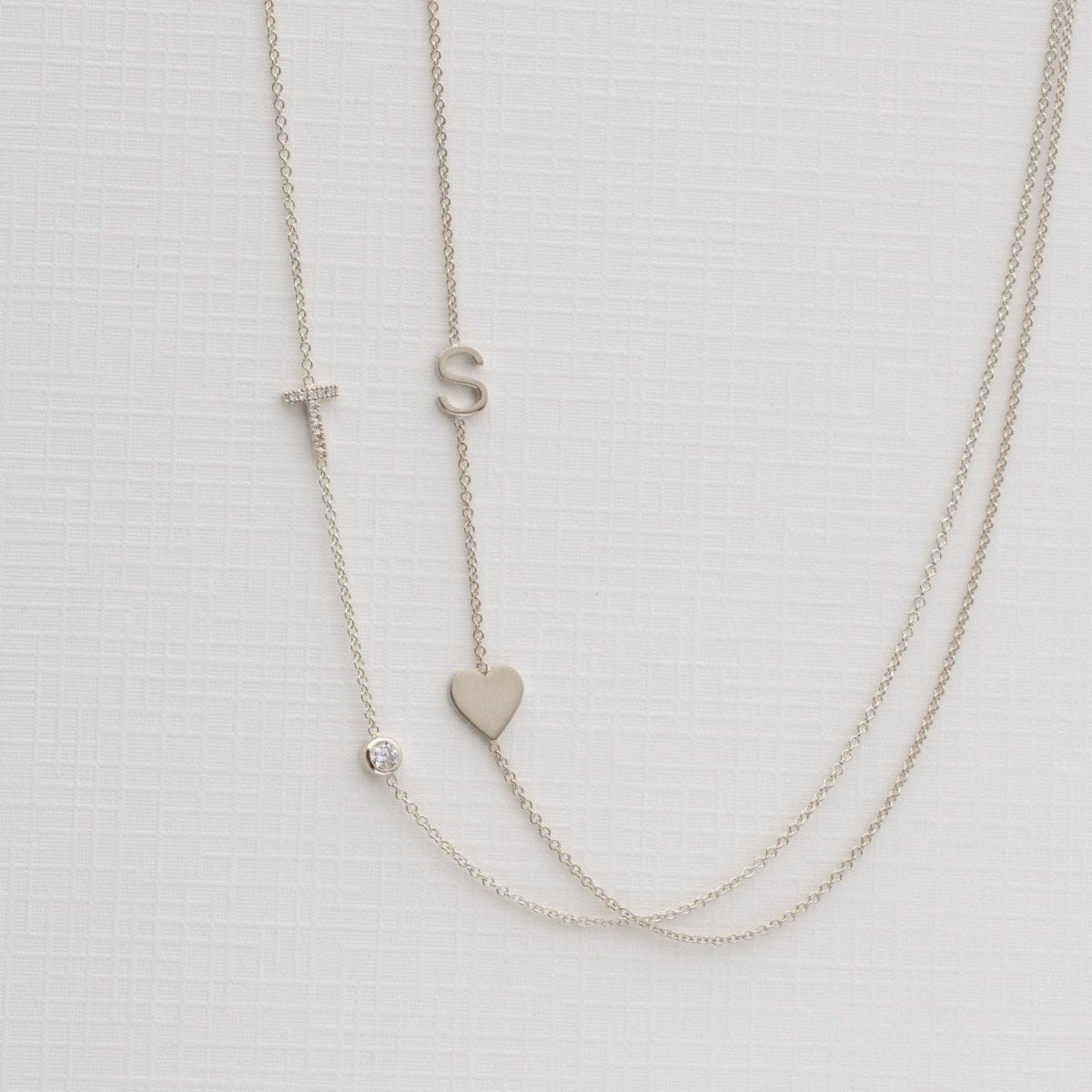 A Monogram Necklace with Heart White Gold and a letter S charm, and a pavé Monogram Necklace with Diamond White Gold and a letter T charm, on a neutral background. 