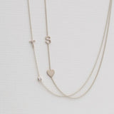 A Monogram Necklace with Heart White Gold and a letter S charm, and a pavé Monogram Necklace with Diamond White Gold and a letter T charm, on a neutral background. 