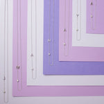 Seven Monogram Necklaces, each with a letter charm (J, M, A, H, A, Y and A) and a diamond charm, laid horizontally against variations of purple coloured paper.”