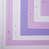 Seven Monogram Necklaces, each with a letter charm (J, M, A, H, A, Y and A) and a diamond charm, laid horizontally against variations of purple coloured paper.”