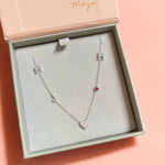 A Custom Gold Necklace in white gold with five charms (two pavé Hs, a pavé J, a pavé heart and a birthstone) in a jewellery box against a pink background.