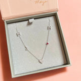 A Custom Gold Necklace in white gold with five charms (two pavé Hs, a pavé J, a pavé heart and a birthstone) in a jewellery box against a pink background.