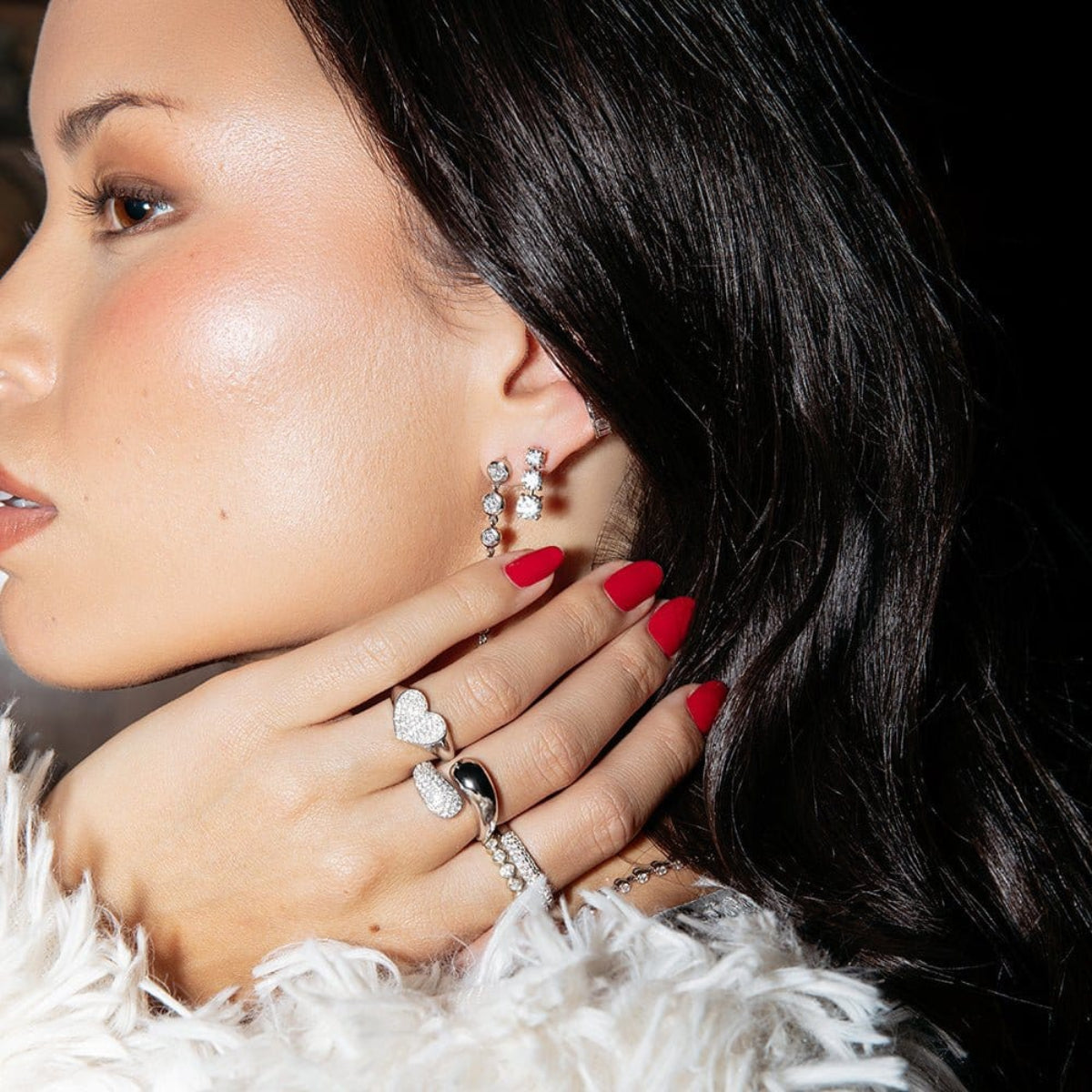 A woman wearing the Evermore Ring, a Cosmo Ring, a Make It a Double Ring, and a Love on the Rocks Ring, along with the Triple Sec Diamond Earring, the Night Cap Ear Cuff, and a Diamond Cascade Earring, all in white gold, with red nails and black hair.