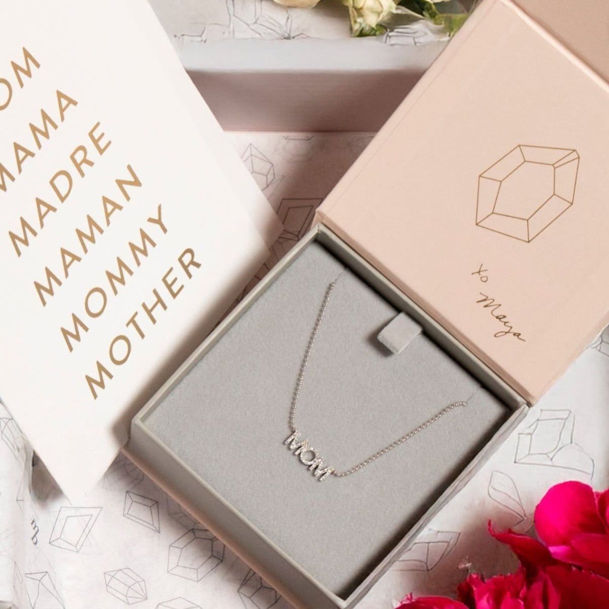 A Pavé MOM Necklace in white gold and a beaded chain displayed in a Maya Brenner jewellery box, accompanied by a paper featuring the words MOM, MAMA, MADRA, MAMAN, MOMMY, and MOTHER.