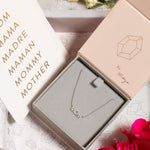 A Pavé MOM Necklace in white gold and a beaded chain displayed in a Maya Brenner jewellery box, accompanied by a paper featuring the words MOM, MAMA, MADRA, MAMAN, MOMMY, and MOTHER.