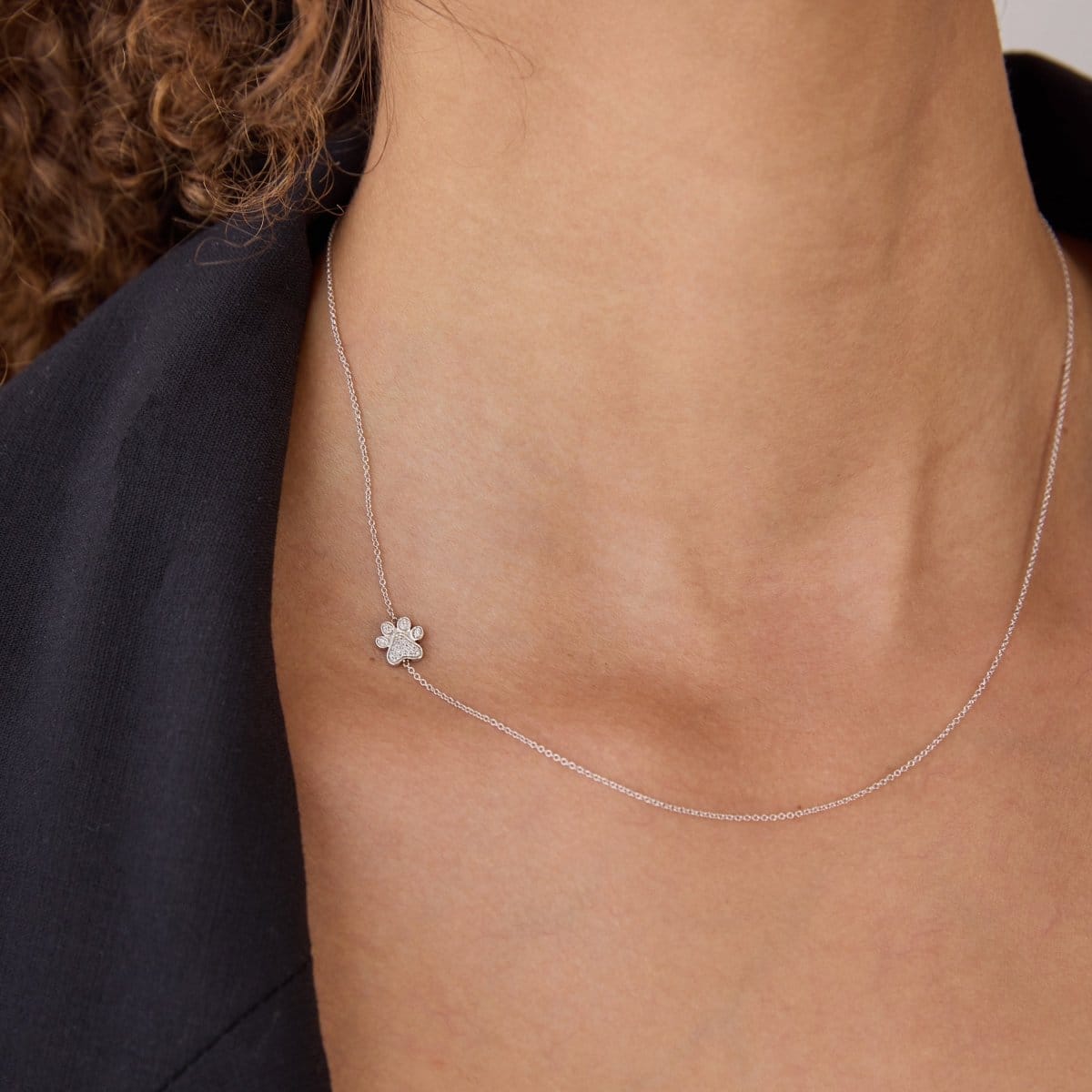 A close-up of a woman's neck wearing a white gold 14k Gold Asymmetrical Charm Necklace - Paw with a cable chain, styled with a black jacket. 