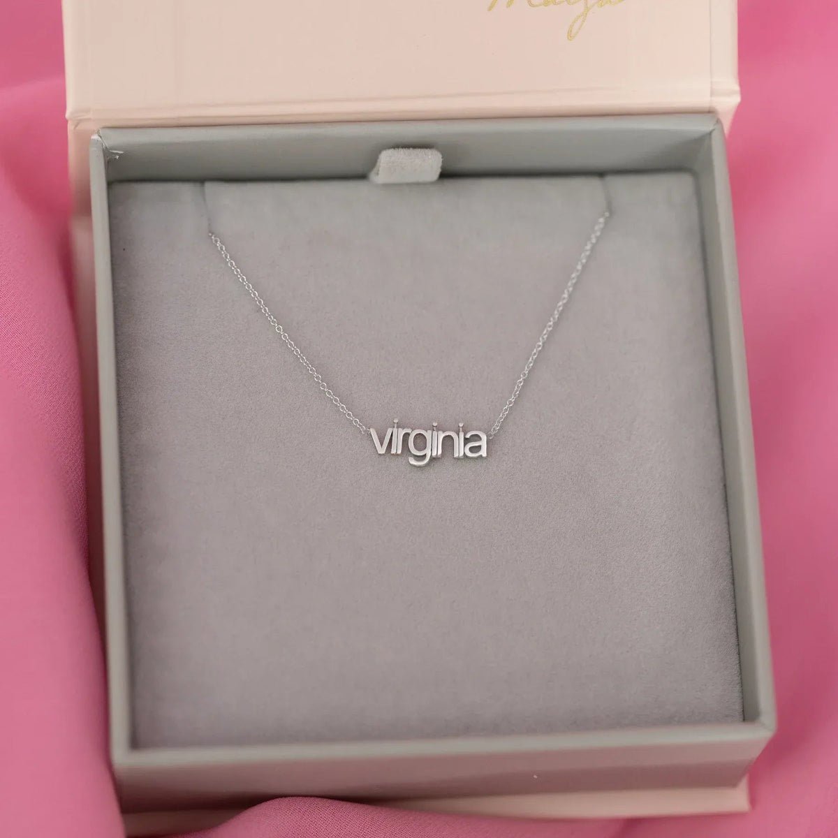 A white gold Petite Nameplate Necklace with 'virginia' in a Maya Brenner jewellery box with a pink background. 