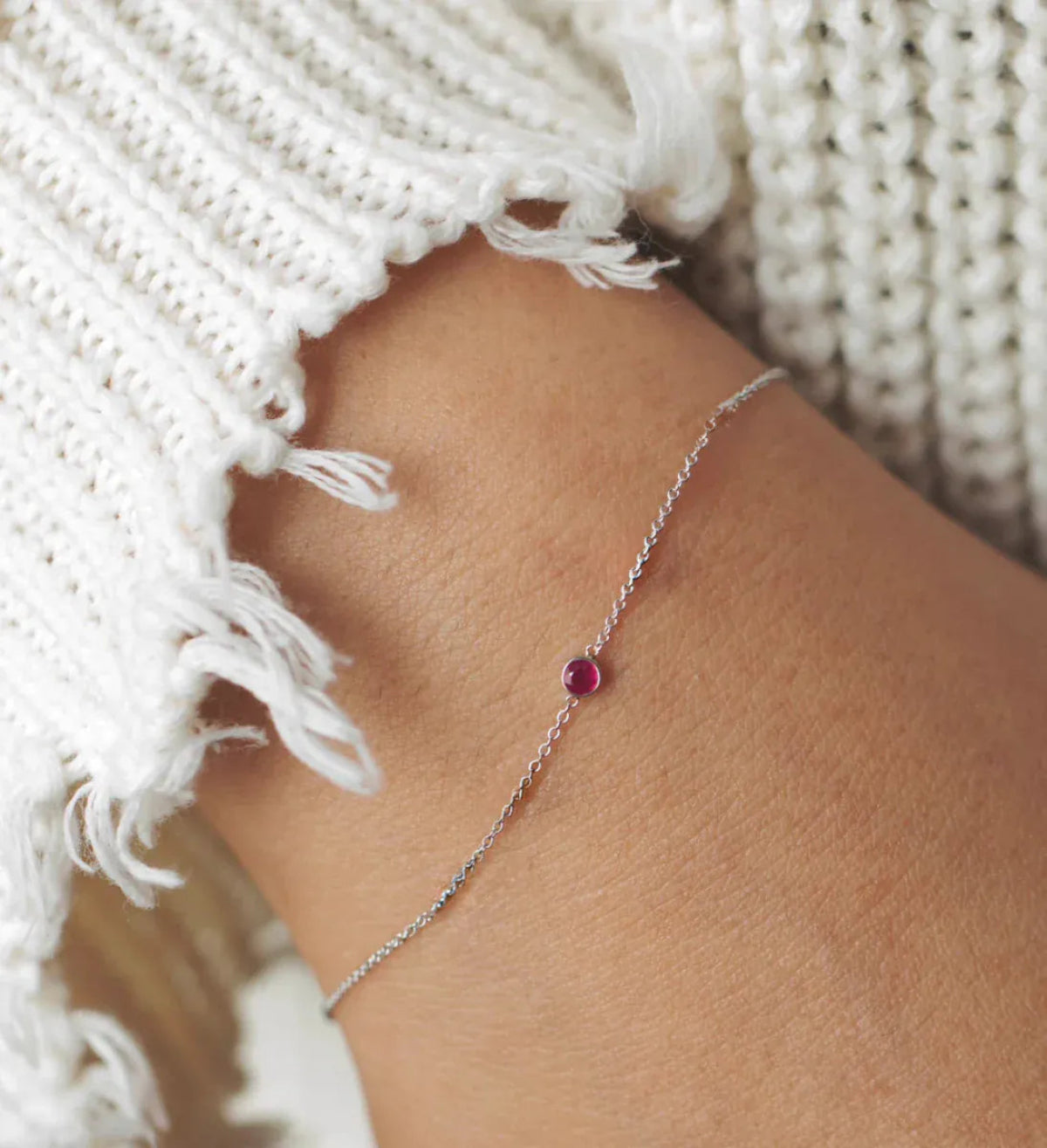 A wrist wearing a white gold Birthstone Bracelet with a ruby birthstone, styled with a white knit.