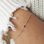 A wrist wearing a white gold Birthstone Bracelet with a ruby birthstone, styled with a white knit.