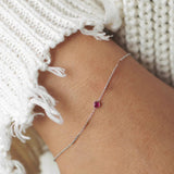 A wrist wearing a white gold Birthstone Bracelet with a ruby birthstone, styled with a white knit.