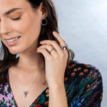A woman wearing a white gold Thick Band, Delicate Beaded Band, 14k Gold Letter Ring, Sharktooth Necklace, Circle Ear Jacket and a Simple Bangle, styled with a colourful patterned top against a light grey background. 