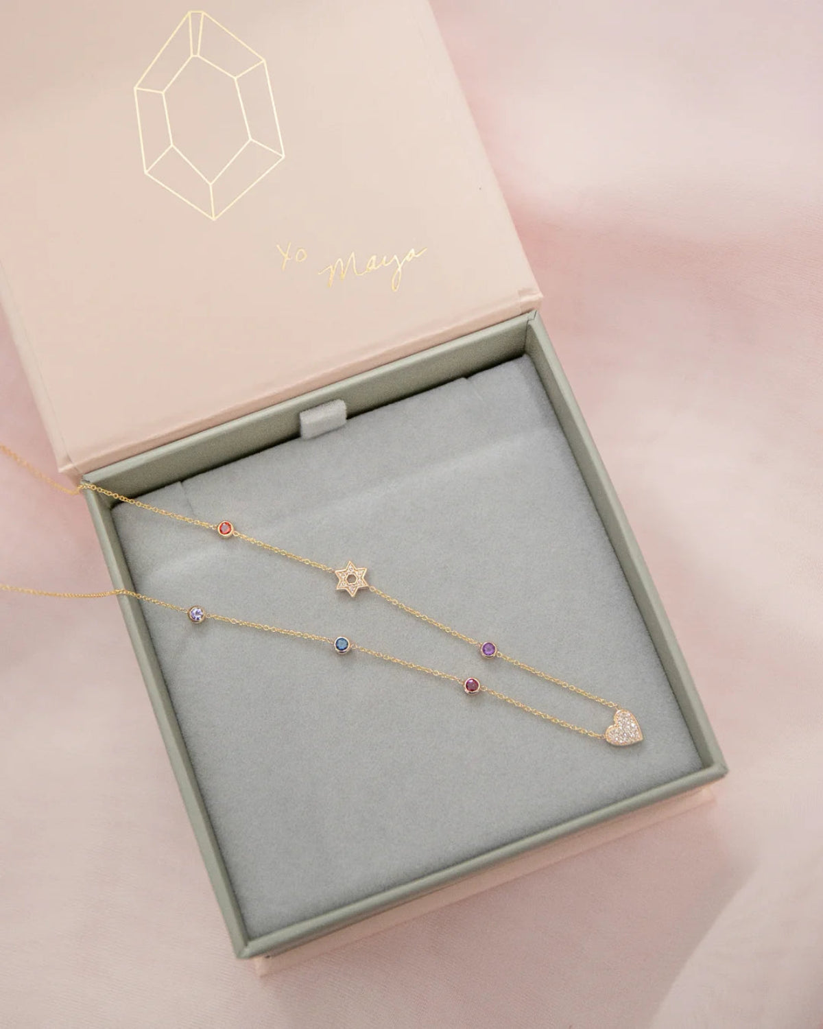 A Custom Gold Necklace in yellow gold with seven charms (five birthstones, a pavé Star of David and a pavé heart) in a Maya Brenner jewellery box against a light pink background.