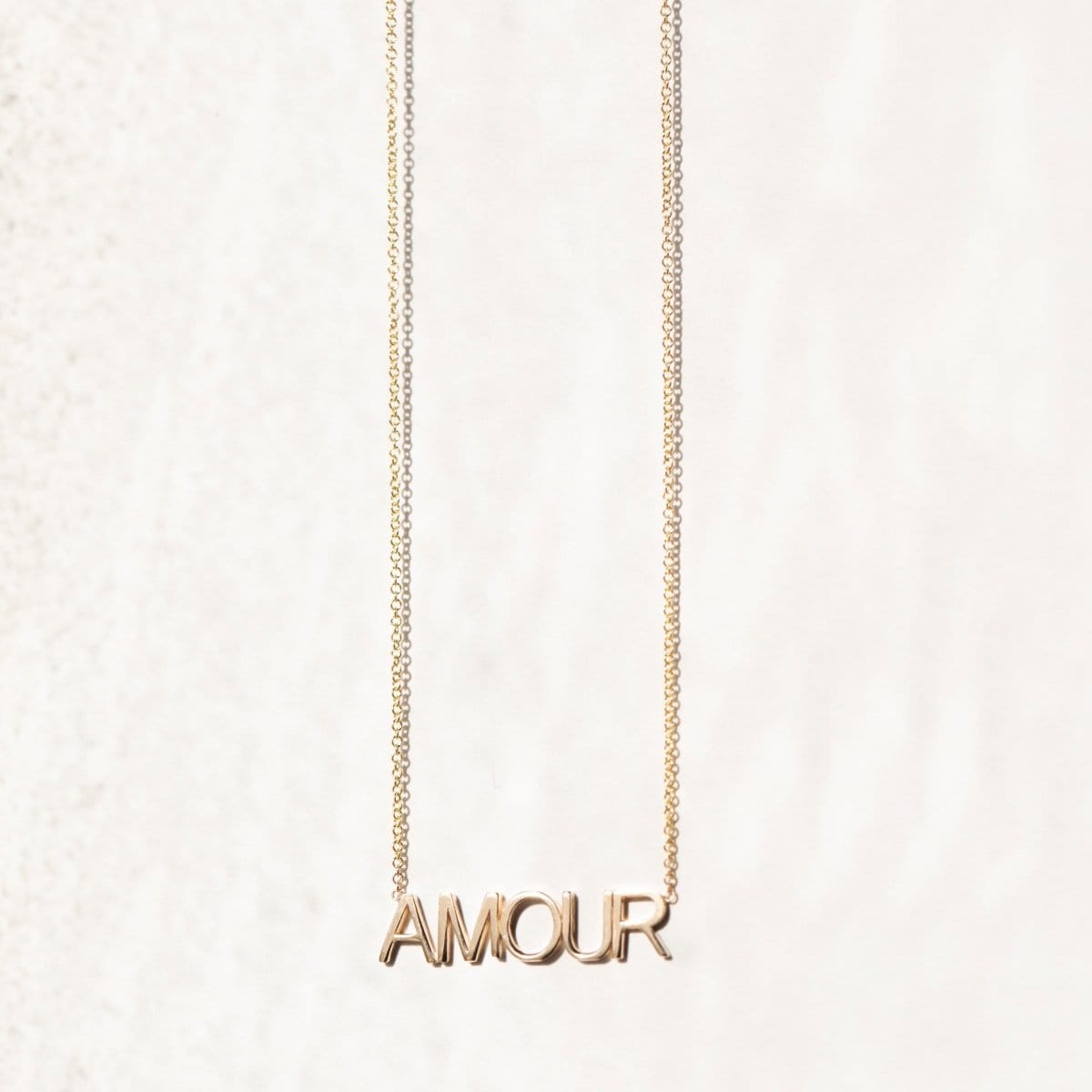 An AMOUR Necklace in yellow gold on a cable chain on an off-white background. 