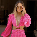 A blonde woman wearing a Zodiac Necklace - Aries, in a bright pink long sleeve dress and sunglasses against a dark background.