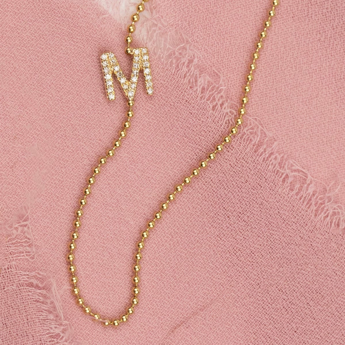 A yellow gold beaded necklace with a pave letter M charm on a pink fabric background.