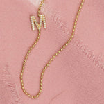 A yellow gold beaded necklace with a pave letter M charm on a pink fabric background.