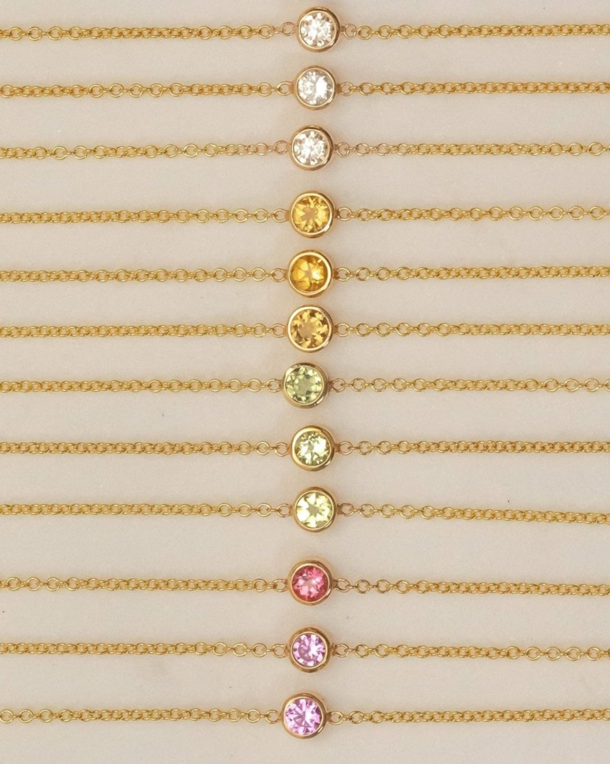 Twelve yellow gold 14K Gold Asymmetrical Birthstone Necklaces - three Diamond (April), three Citrine (November), three Peridot (August), one Garnet (January) and two Pink Tourmaline (October) laid against a neutral background. 