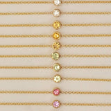 Twelve yellow gold 14K Gold Asymmetrical Birthstone Necklaces - three Diamond (April), three Citrine (November), three Peridot (August), one Garnet (January) and two Pink Tourmaline (October) laid against a neutral background. 