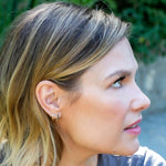 A woman wearing an Alli Webb Faceted Huggie and Onyx Huggie with blonde hair against a blurred greenery background. 