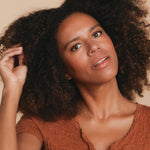 A woman with dark curly hair wearing a rust top and a Custom Gold Necklace in yellow gold with three charms, two birthstones on the left side and a cross charm on the right, against a beige background.