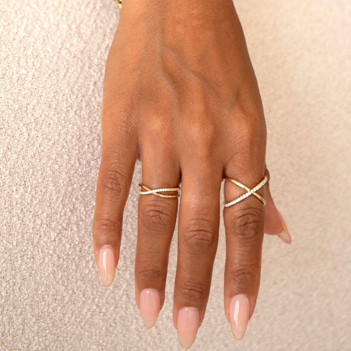 A hand wearing a yellow gold Criss-Cross Diamond Duo Ring and Criss-Cross Diamond Daze Ring against a cream background. 