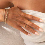 A woman's hand wearing a Criss-Cross Duo Ring, Rolo Bracelet, an Element Short Link Bracelet and a Down the Line Bracelet, in front of her white strapless against against a cream fabric background. 