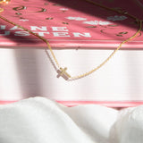 A 14k Gold Asymmetrical Charm Necklace - Cross in yellow gold and cable chain, laid on top of a book with a dark pink cover on a white fabric surface.