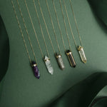 Five The Retreat Necklaces (Healing, Clarity, Protection, Courage and Happiness) laid against a dark green background.
