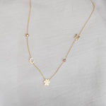 A Custom Gold Necklace in yellow gold with five charms, two birthstones, the letters C and M, and a paw, on a neutral background.