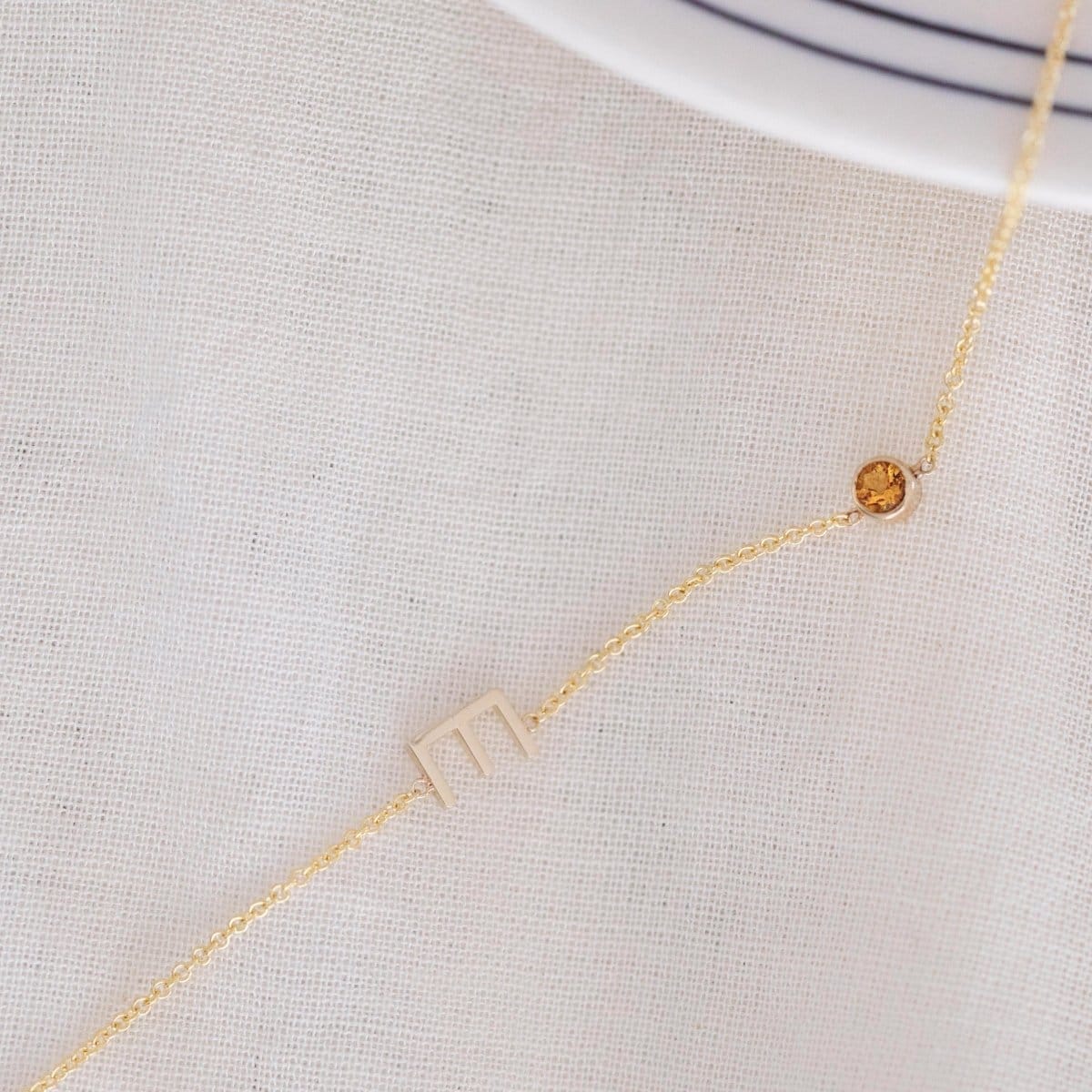A Custom Gold Necklace in yellow gold with a letter E charm and a birthstone, on an off-white fabric background. 
