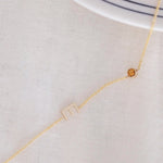 A Custom Gold Necklace in yellow gold with a letter E charm and a birthstone, on an off-white fabric background. 
