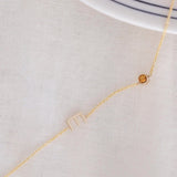 A Custom Gold Necklace in yellow gold with a letter E charm and a birthstone, on an off-white fabric background. 