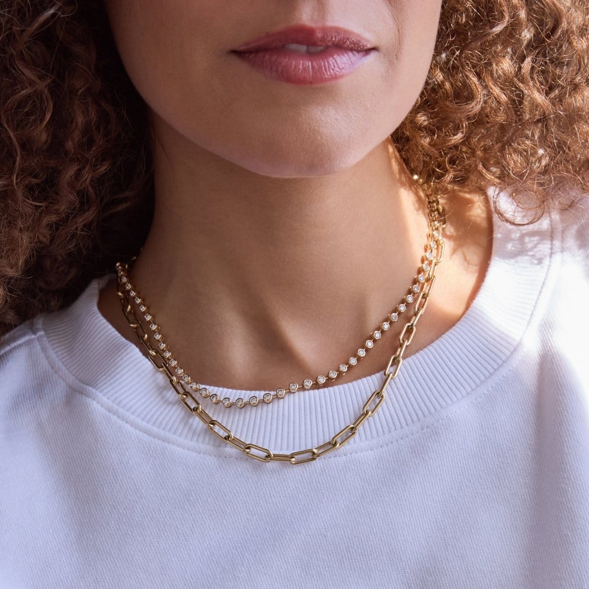 Close-up of a person's neck wearing The Ultimate Diamond Necklace and a Bold Link Chain, with a white t-shirt and brown curly hair. 