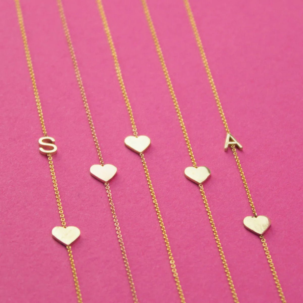 Five yellow gold necklaces laid out against a bright pink background, two Monogram Necklaces with Heart Yellow Gold with the letters S and A and three heart charm necklaces.