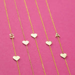 Five yellow gold necklaces laid out against a bright pink background, two Monogram Necklaces with Heart Yellow Gold with the letters S and A and three heart charm necklaces.