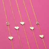 Five yellow gold necklaces laid out against a bright pink background, two Monogram Necklaces with Heart Yellow Gold with the letters S and A and three heart charm necklaces.