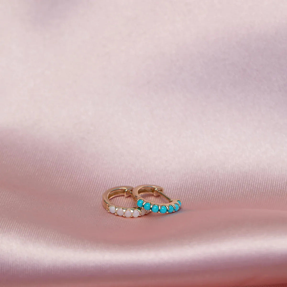 An Alli Webb Opal Huggie and Turquoise Huggie on a light pink silk fabric. 