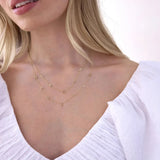 A close-up of a woman’s neck area wearing two Custom Gold Necklaces in yellow gold, one with five letter charms (S, A, D, I and E) and the other with seven letter charms (M, A, D, E, L, Y and N), with blonde hair and a white top against a white background.