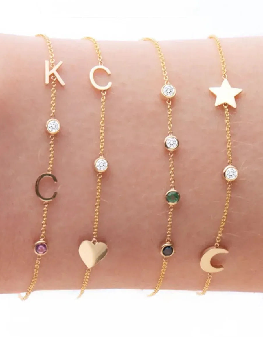 A wrist wearing four yellow gold Custom Gold Bracelets: two with three charms (C, a diamond birthstone and a heart, and a star, a diamond birthstone and a moon) and two with four charms (K, C, diamond and amethyst birthstones, and two diamonds, one emerald and one sapphire), on a white background.