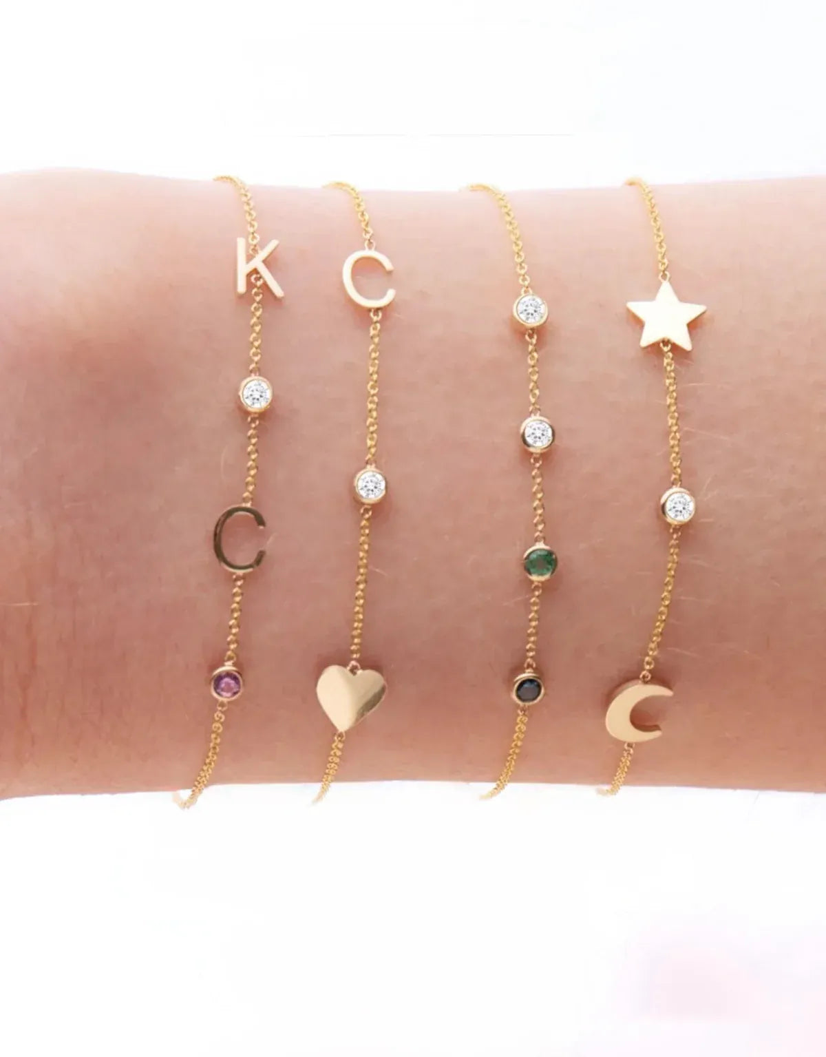A wrist wearing four yellow gold Custom Gold Bracelets: two with three charms (C, a diamond birthstone and a heart, and a star, a diamond birthstone and a moon) and two with four charms (K, C, diamond and amethyst birthstones, and two diamonds, one emerald and one sapphire), on a white background.