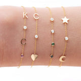 A wrist wearing four yellow gold Custom Gold Bracelets: two with three charms (C, a diamond birthstone and a heart, and a star, a diamond birthstone and a moon) and two with four charms (K, C, diamond and amethyst birthstones, and two diamonds, one emerald and one sapphire), on a white background.