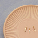 Three Gold Letter Charms in I, L and A on a textured light orange plate with a light grey background. 