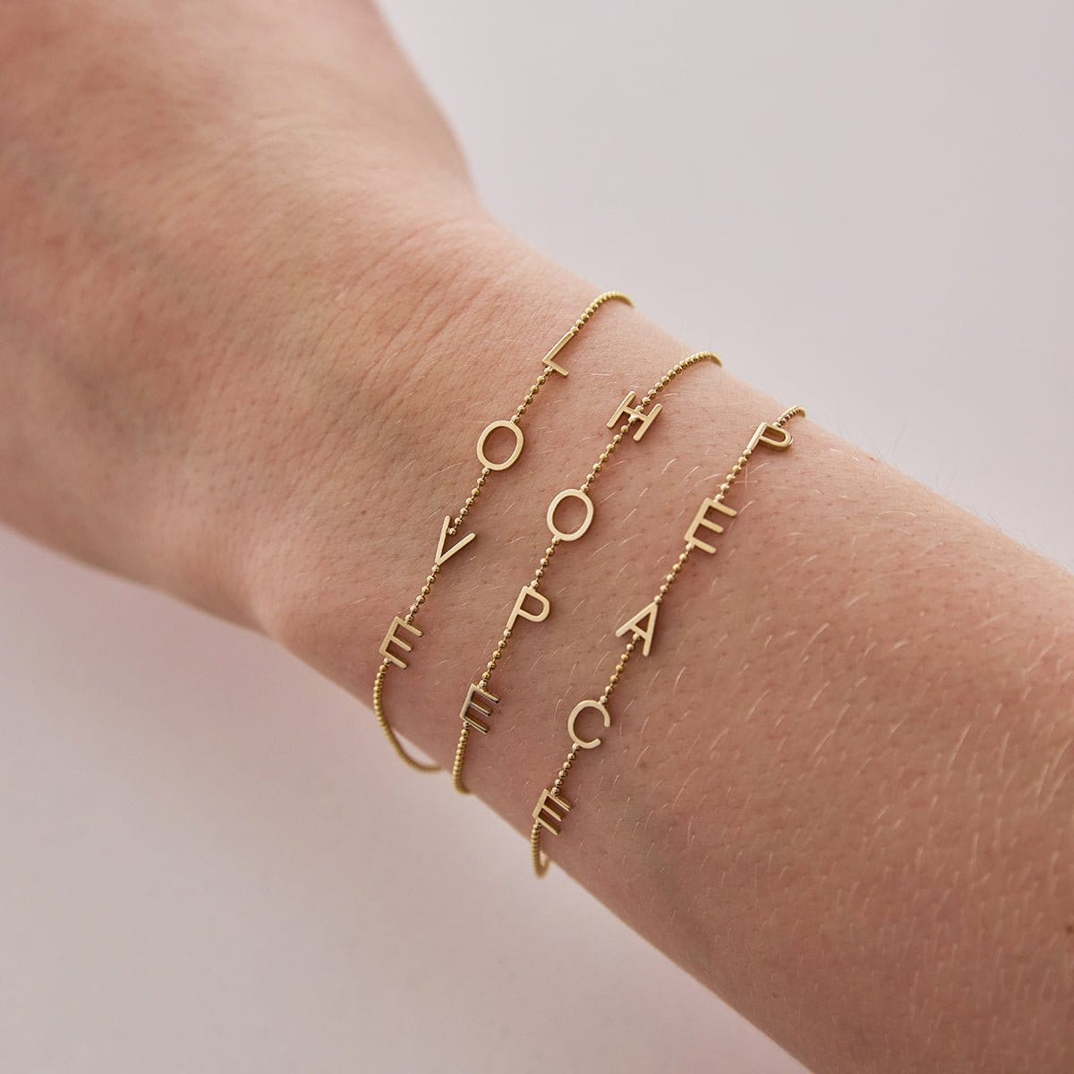 A wrist wearing two Custom Gold Bracelets – 4 Letters (L, O, V, E and H, O, P, E) and one 5 Letters (P, E, A, C, E) in bead chains, on a neutral background.