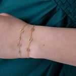 A close-up of a wrist wearing two yellow gold Custom Gold Bracelets in beaded chains, one with four letters M, A, M, A and the other with three letters M, U, M, against a dark turquoise blue silk fabric.