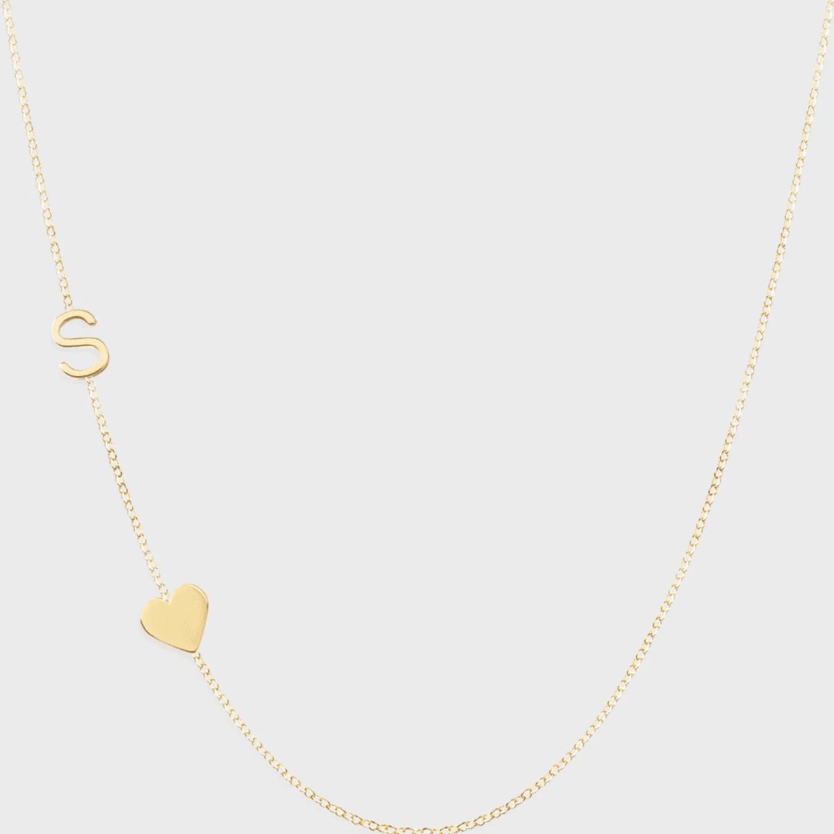 A Monogram Necklace with Heart Yellow Gold and a letter S charm on a light grey background. 