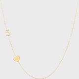 A Monogram Necklace with Heart Yellow Gold and a letter S charm on a light grey background. 