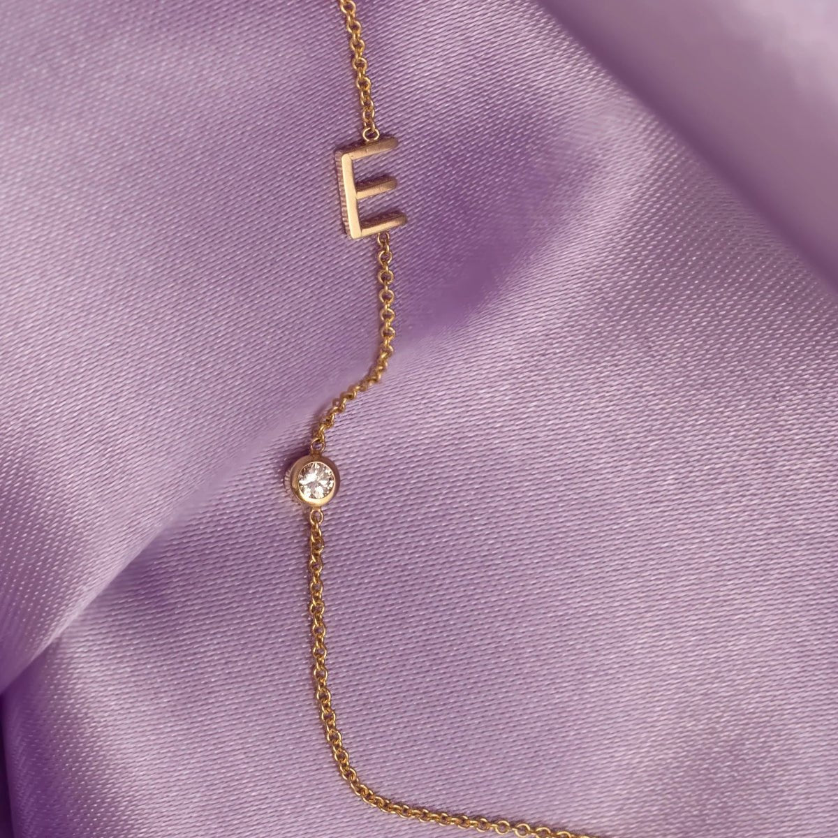 A Monogram Necklace with Diamond and the letter E in yellow gold, against light purple silk fabric as the background.
