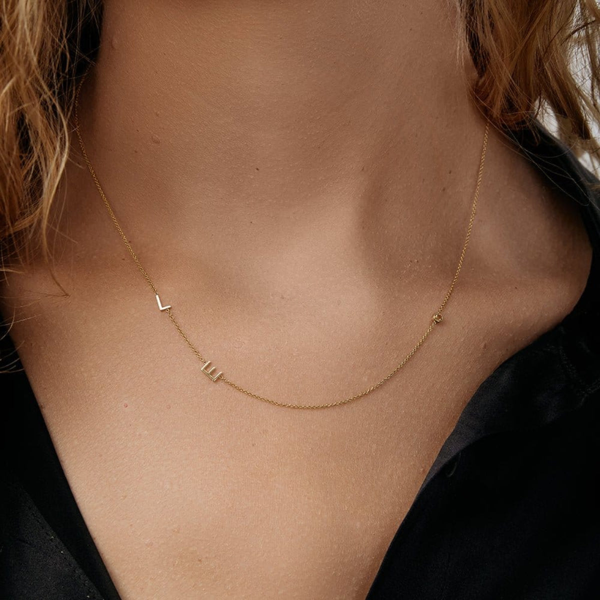 A close-up of a woman’s neck wearing a Custom Gold Necklace in yellow gold with the letters L and E on a left side of the necklace and a birthstone on the right, and a black shirt. 
