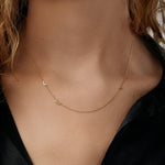 A close-up of a woman’s neck wearing a Custom Gold Necklace in yellow gold with the letters L and E on a left side of the necklace and a birthstone on the right, and a black shirt. 