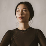 A woman in a black sheer long sleeve top wearing a yellow gold necklace with a letter S charm and drop earrings with circles.
